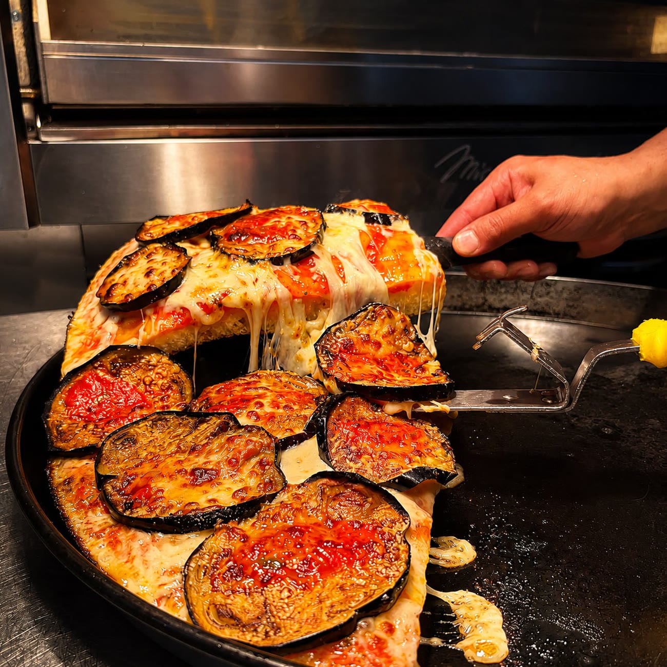 Trancio di pizza alla parmigiana sollevato dalla teglia con il tiraggio della mozzarella davanti al forno